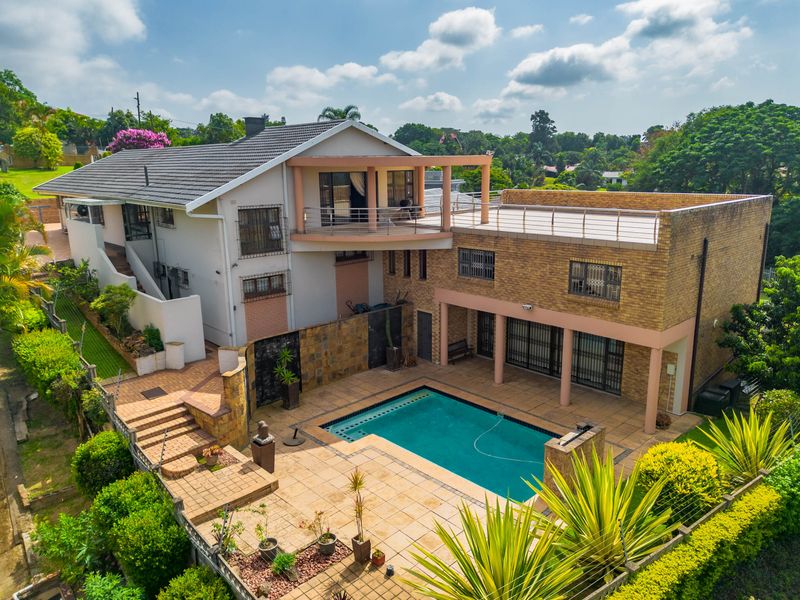 Aerial view of a multi-level residential property featuring a swimming pool, expansive tiled patio area, upper-level balcony, and established garden surroundings.