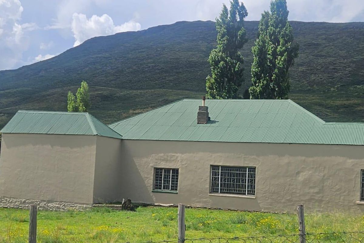 Rear view of a farm cottage featuring a green corrugated metal roof, stone foundation base, and security-barred windows set against a mountain backdrop.
