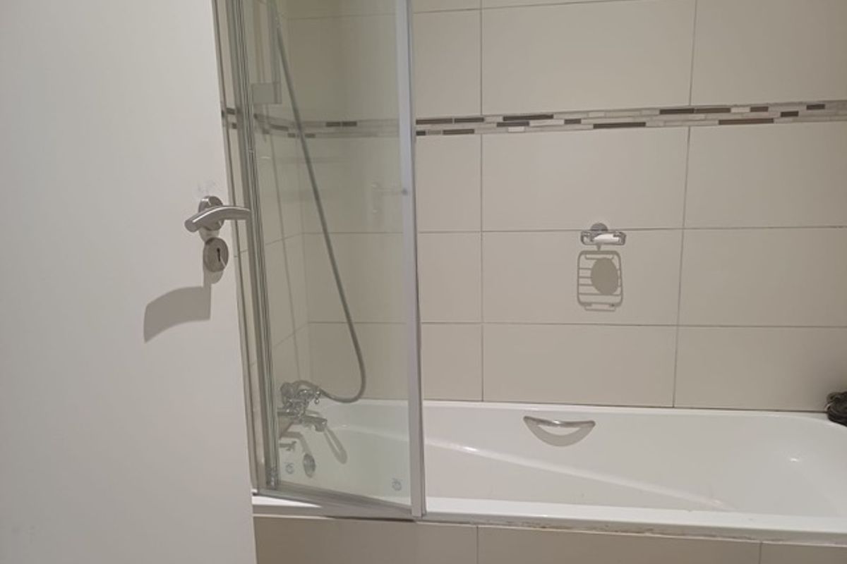A functional bathroom featuring a built-in bathtub with a glass shower screen and neutral wall tiling with a decorative accent border.