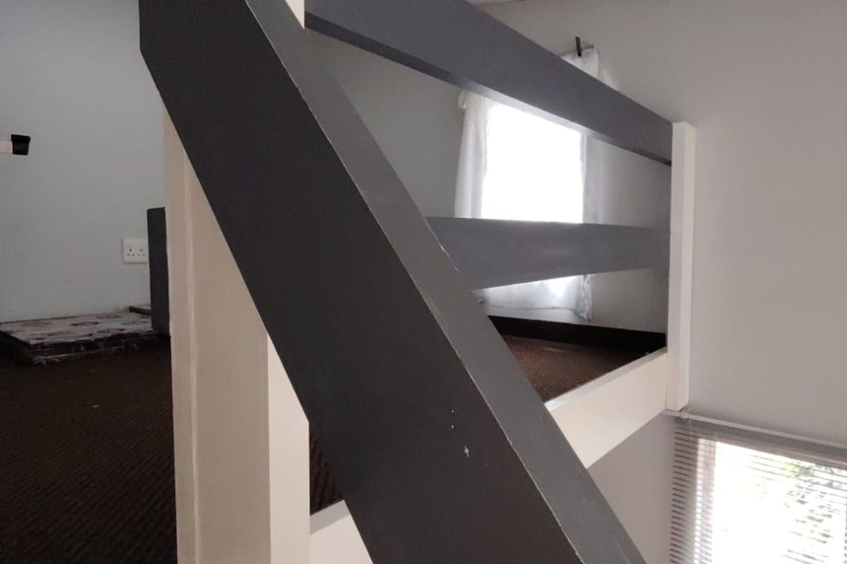 A mezzanine level bedroom area featuring a dark painted staircase railing, carpeted flooring, and natural light from a window with venetian blinds.