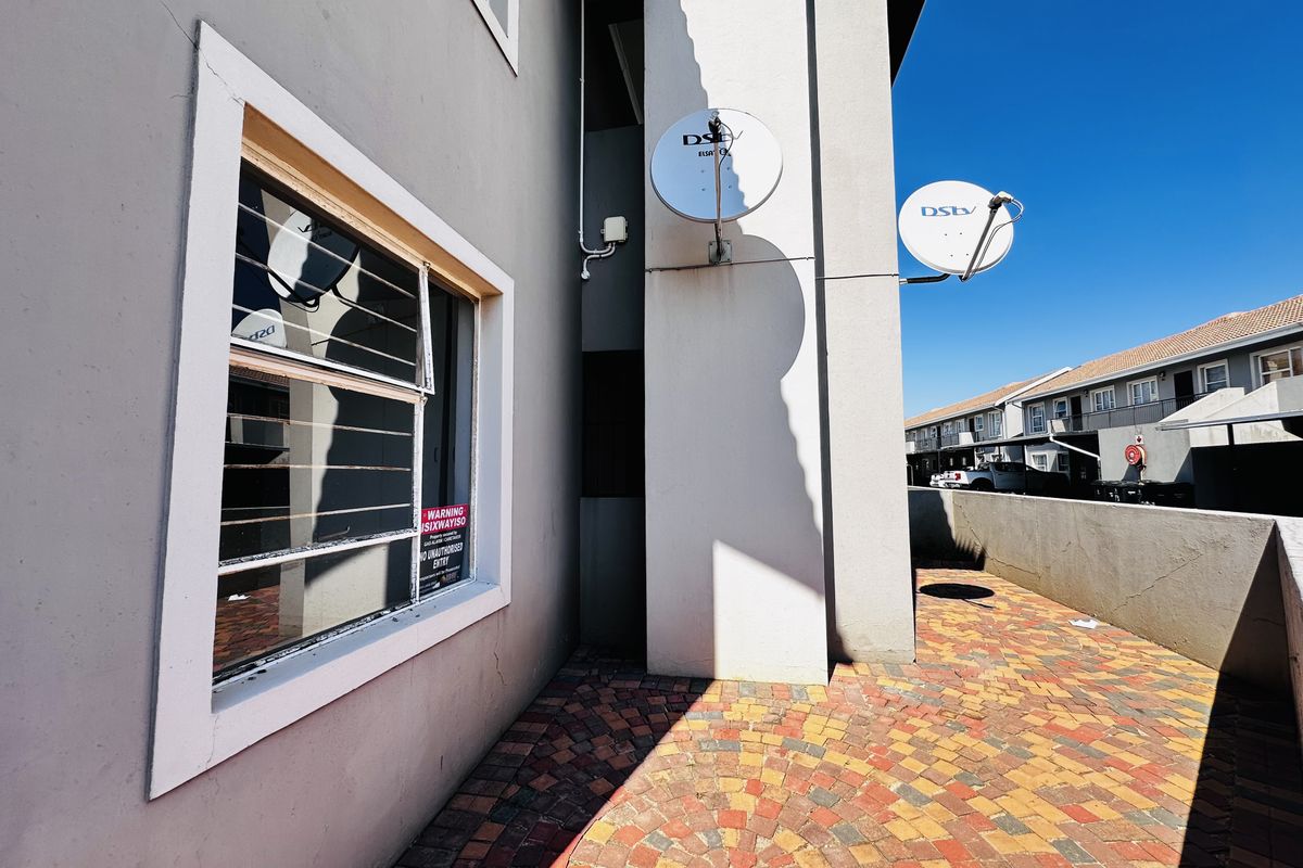 Rear exterior view of the apartment featuring a paved patio area, security bars on the window, and mounted satellite dishes.