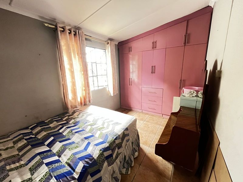 A bedroom featuring built-in pink cupboards, tiled flooring, and a window with security bars, offering a functional space with potential for cosmetic updates.