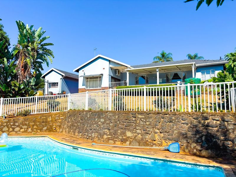 A well-maintained outdoor pool area featuring a stone retaining wall, white perimeter fencing, and a covered patio overlooking the swimming pool.