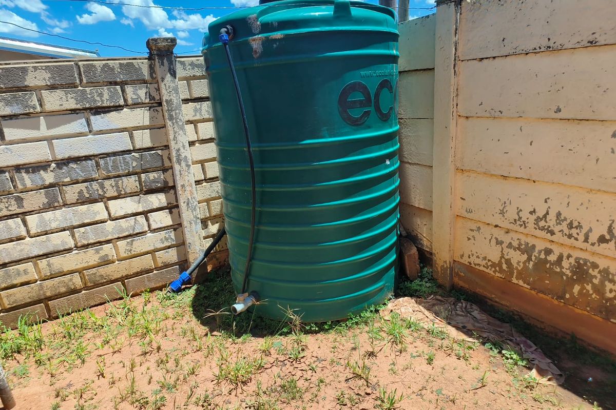 An outdoor area featuring a large green plastic water storage tank positioned against a weathered brick and concrete boundary wall.