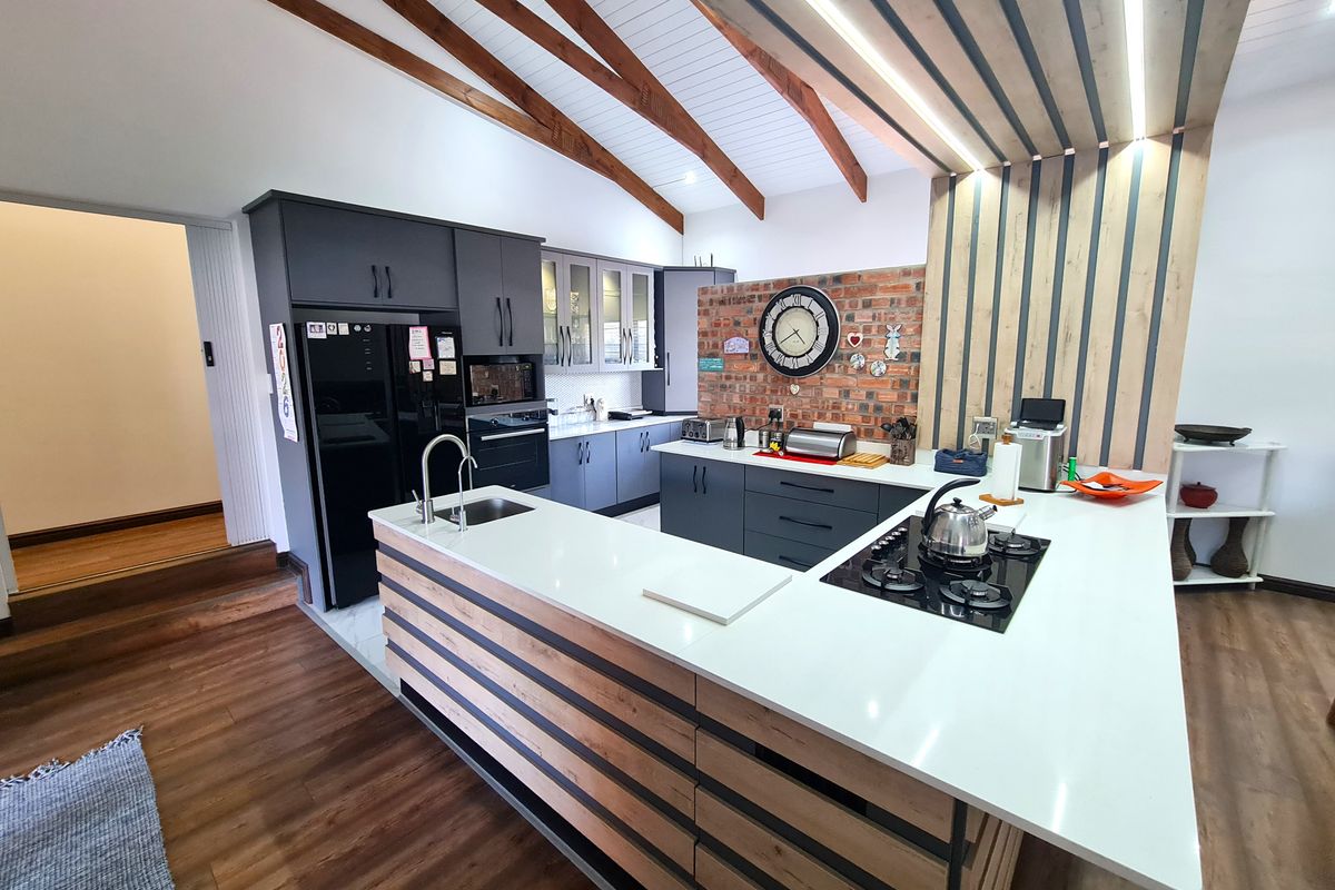 Modern kitchen featuring a large island with stone countertops, gas hob, exposed timber beams, and a striking brick feature wall.