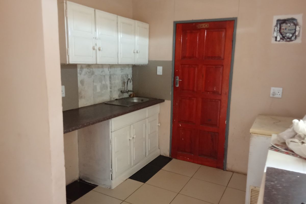 A compact kitchen area featuring basic white cabinetry, a stainless steel sink, tiled flooring, and a prominent red wooden door.