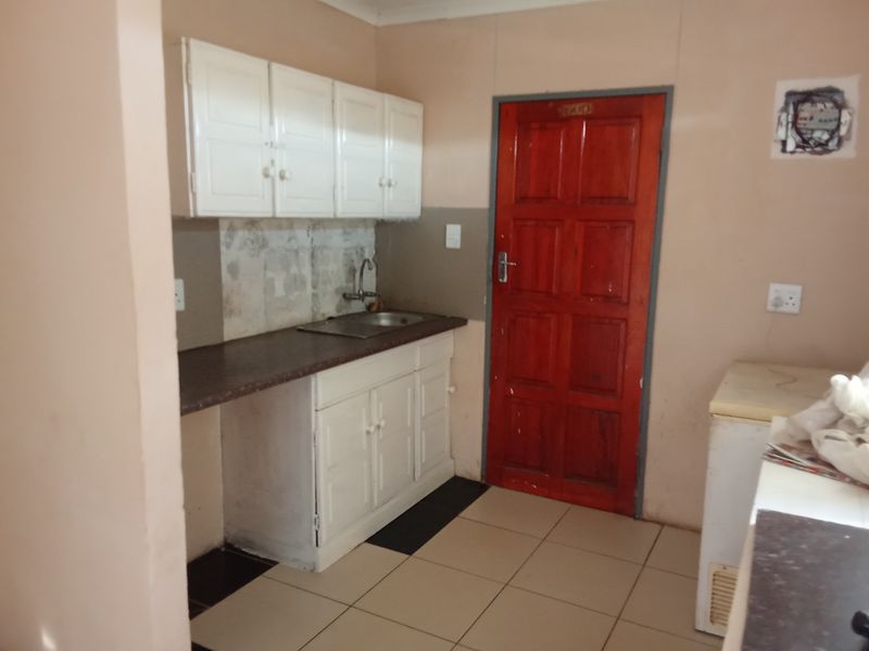 A compact kitchen area featuring basic white cabinetry, a stainless steel sink, tiled flooring, and a prominent red wooden door.