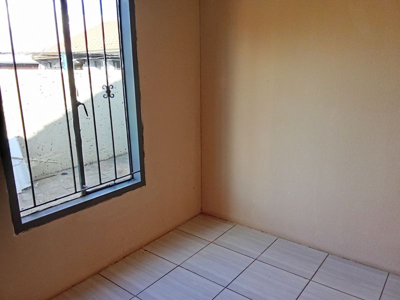 An empty bedroom featuring neutral tiled flooring and a window fitted with security bars, offering a blank canvas for personalization.
