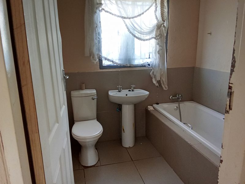 A functional bathroom featuring a standard bathtub, pedestal basin, and toilet, with neutral wall tiling and a window for natural light.