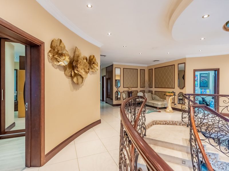 Spacious upper-level hallway featuring elegant wrought iron staircase railings, polished tile flooring, and decorative wall paneling with recessed ceiling lighting.