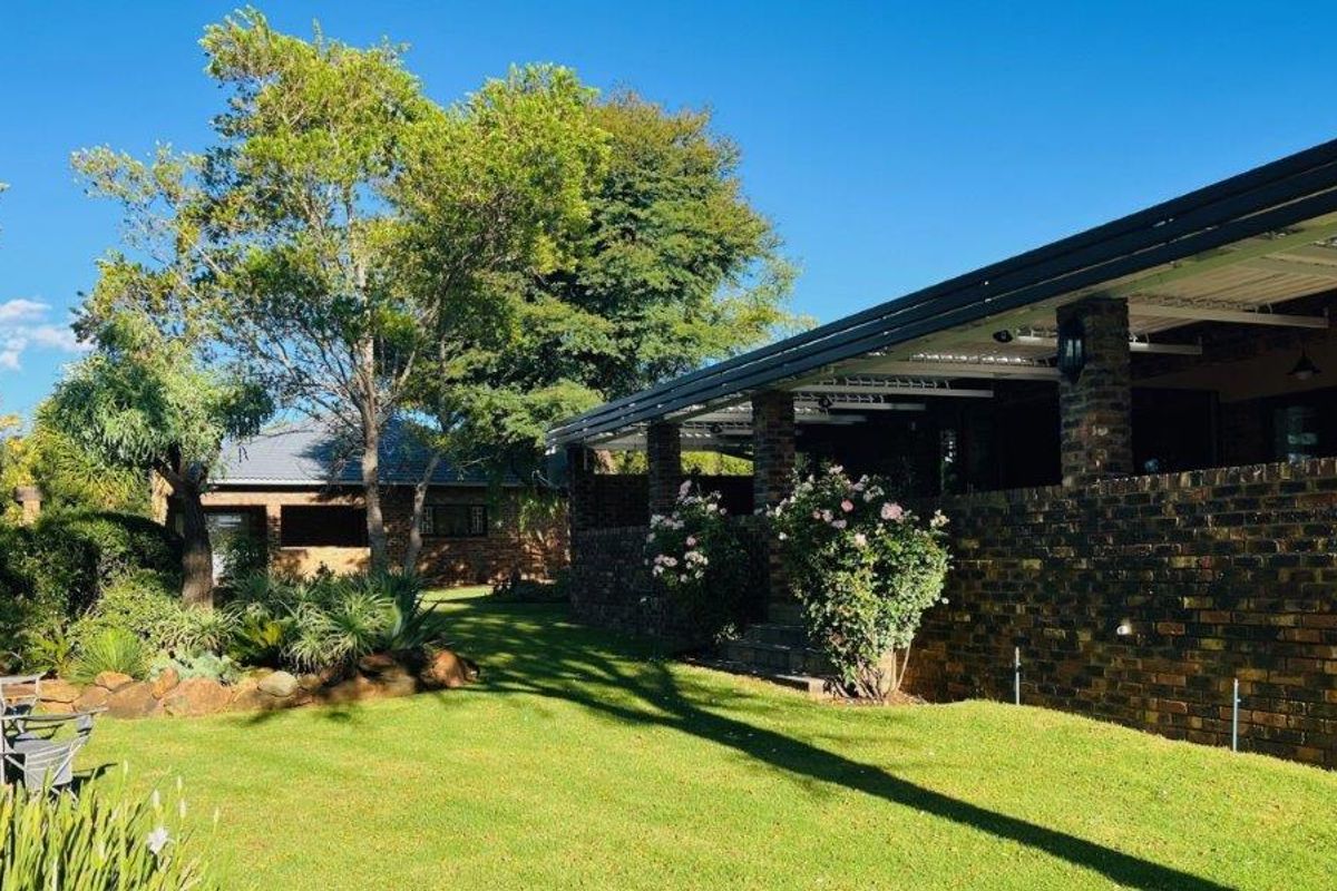 A view of the rear garden featuring a well-maintained lawn, mature trees, and a covered brick patio area attached to the main house.