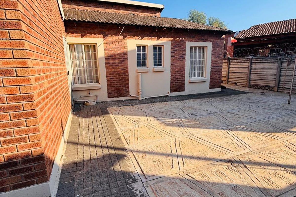 A rear exterior view of the house featuring a paved courtyard area, brick facade, and security fencing along the perimeter.