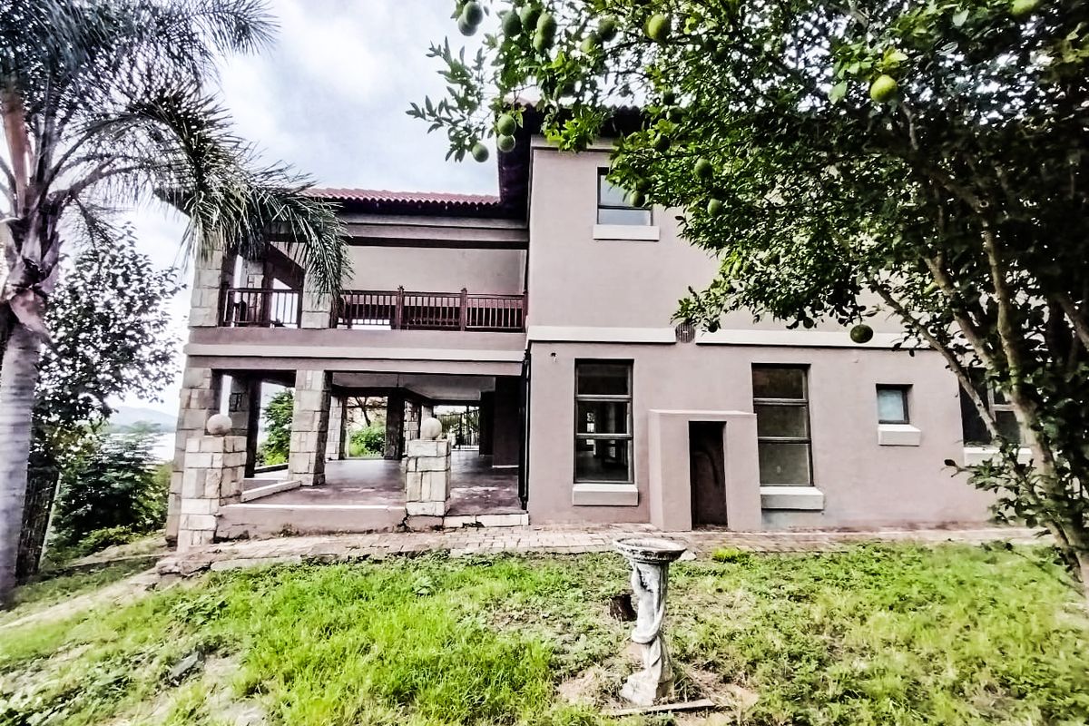 Rear view of a multi-story house featuring a covered patio, upper-level balcony, stone-clad pillars, and a paved walkway leading to the garden.
