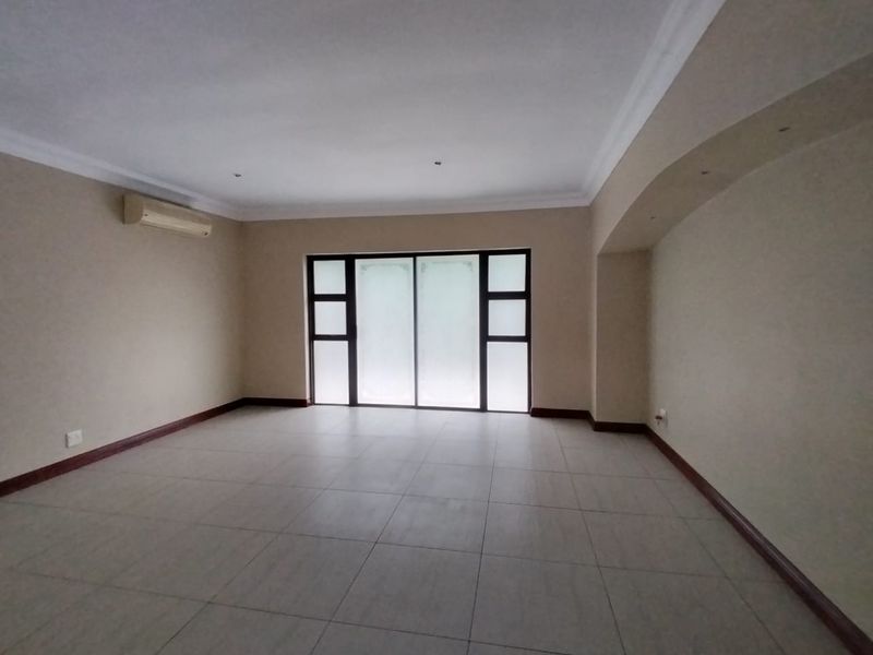 A spacious, empty living room featuring neutral tiled flooring, a wall-mounted air conditioner, and a large sliding glass door providing natural light.