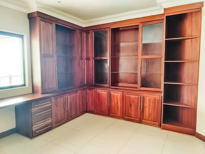 A spacious home office featuring extensive custom-built wooden cabinetry, integrated shelving, glass display units, and a built-in desk area with tiled flooring.