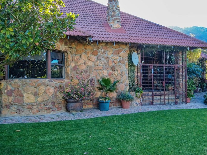 A rear view of the house featuring rustic stone cladding, a tiled roof, a stone chimney, and a paved walkway leading to a secure entrance.