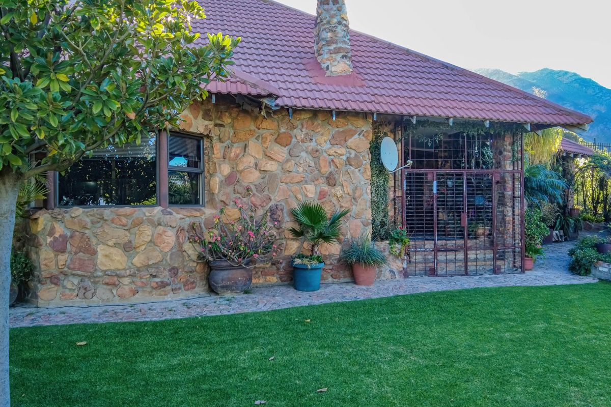 A rear view of the house featuring rustic stone cladding, a tiled roof, a stone chimney, and a paved walkway leading to a secure entrance.