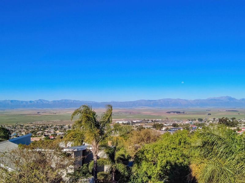 A panoramic elevated view overlooking the surrounding valley, residential area, and distant mountain range under a clear blue sky.