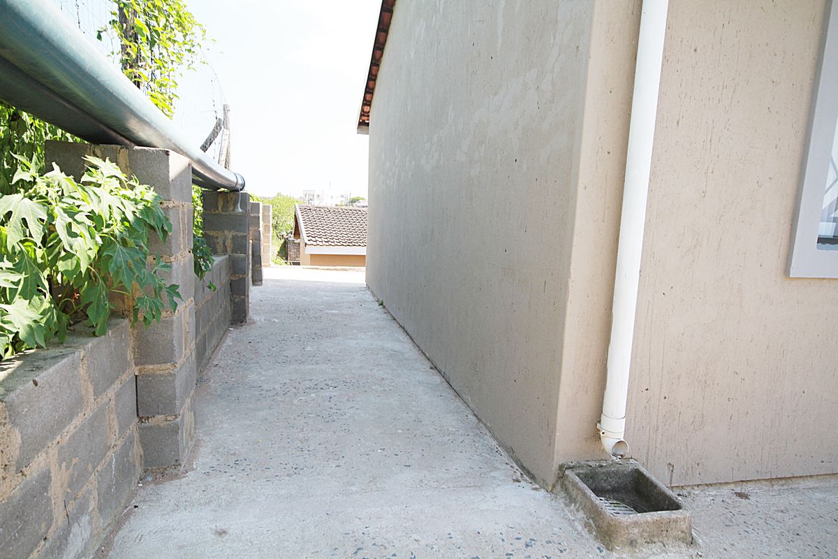 A narrow concrete side passage along the exterior of the house, featuring a downpipe drainage system and a low block retaining wall.