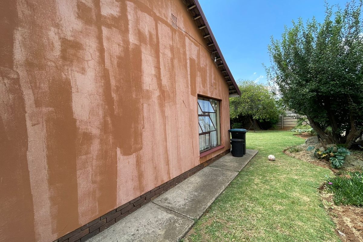 Rear exterior view of the house featuring a concrete pathway, brick plinth, and steel-framed window, with visible uneven paintwork on the exterior wall.