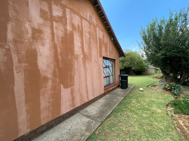 Rear exterior view of the house featuring a concrete pathway, brick plinth, and steel-framed window, with visible uneven paintwork on the exterior wall.