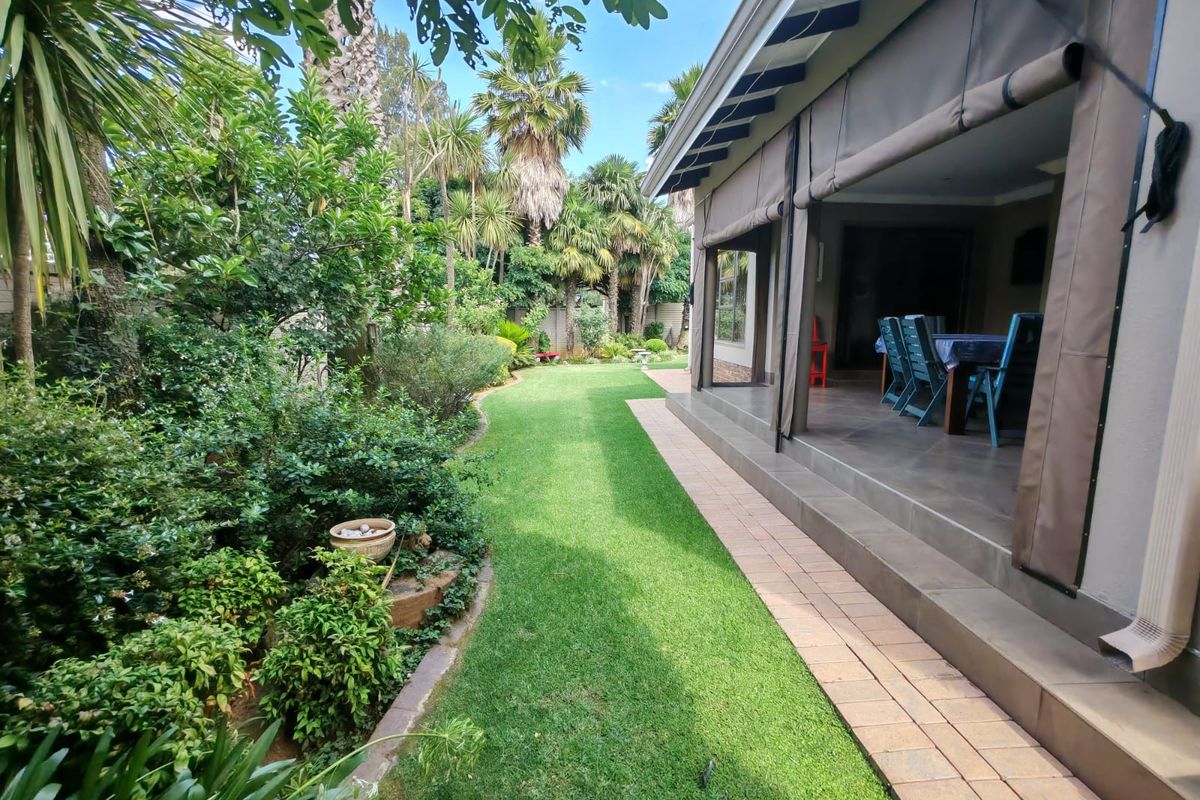 A well-maintained rear garden featuring a lush lawn, mature landscaping, and a spacious covered patio with tiled flooring and retractable privacy blinds.