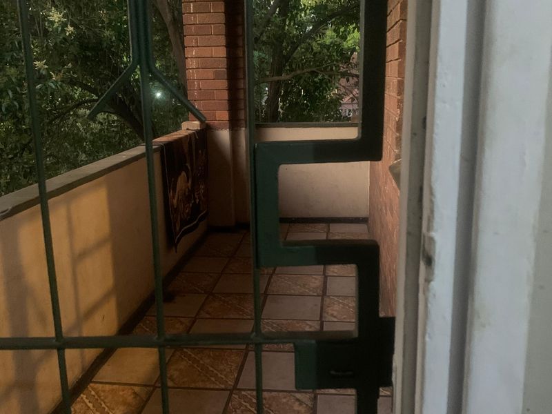 A covered balcony featuring tiled flooring, brick pillars, and a security gate, offering an outdoor space with a view of surrounding trees.