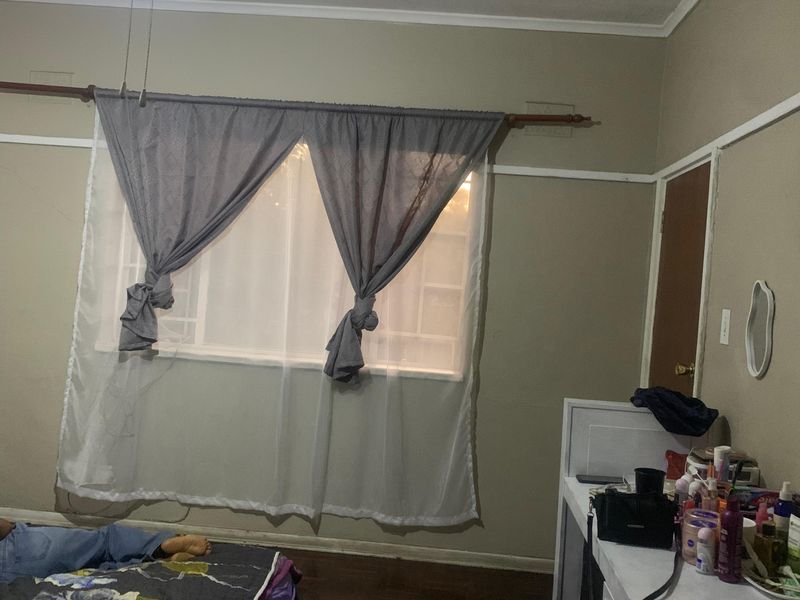 A bedroom featuring parquet flooring, a ceiling fan, and a window. The room shows signs of wear including wall cracks and ceiling staining.