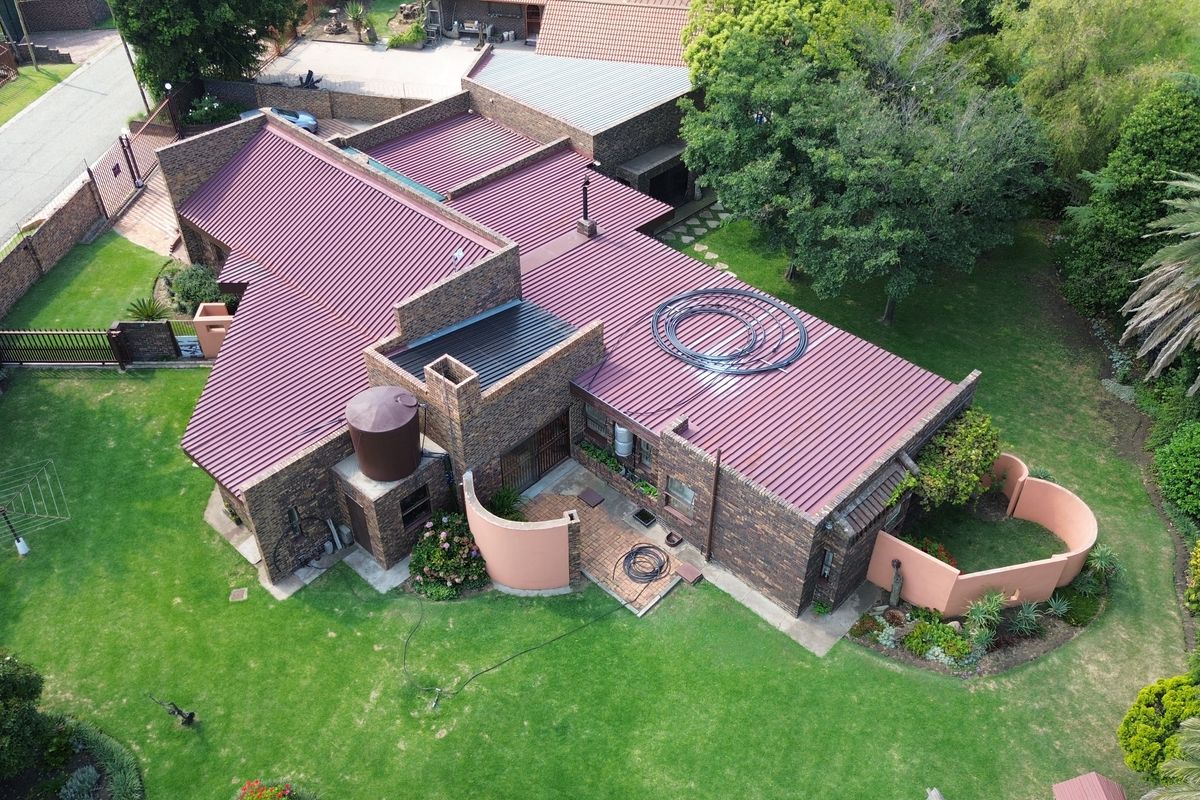 Aerial view of a brick house featuring a corrugated metal roof, solar pool heating system, water storage tank, and established garden surroundings.