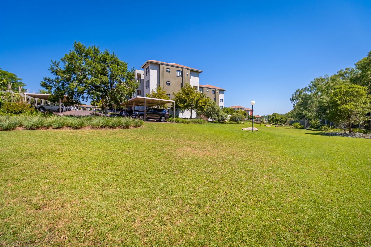 A view of the apartment complex grounds featuring a well-maintained lawn, mature trees, and covered parking structures.