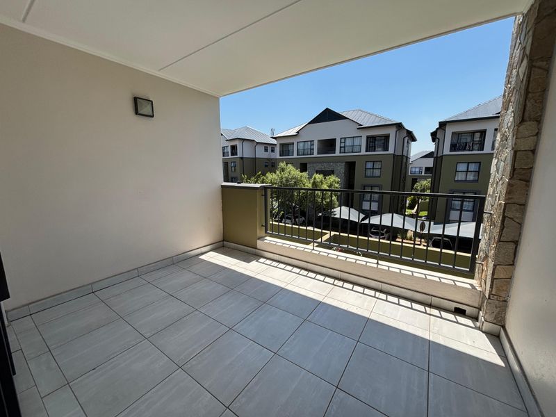 A clean, covered balcony featuring tiled flooring, a metal balustrade, and a decorative stone-clad wall, offering an open view of the apartment complex.