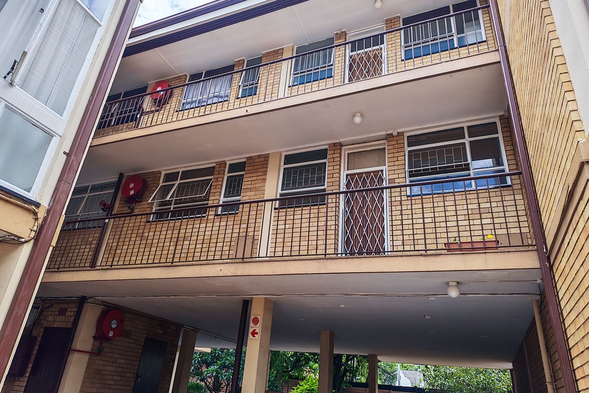 Exterior view of a multi-story apartment building featuring brick facade, open balconies with security gates, and a covered ground-level parking area.