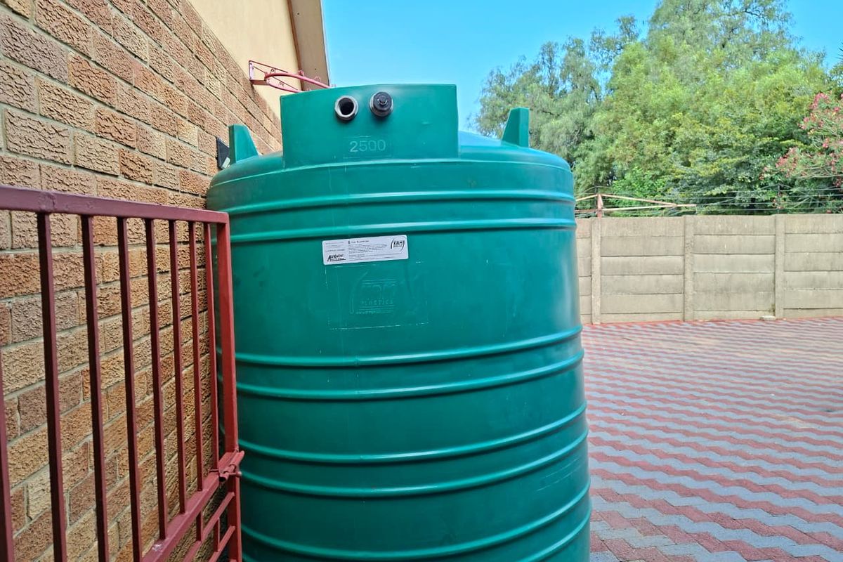 A large 2500-liter green water storage tank installed on a paved exterior area adjacent to the brick building wall.