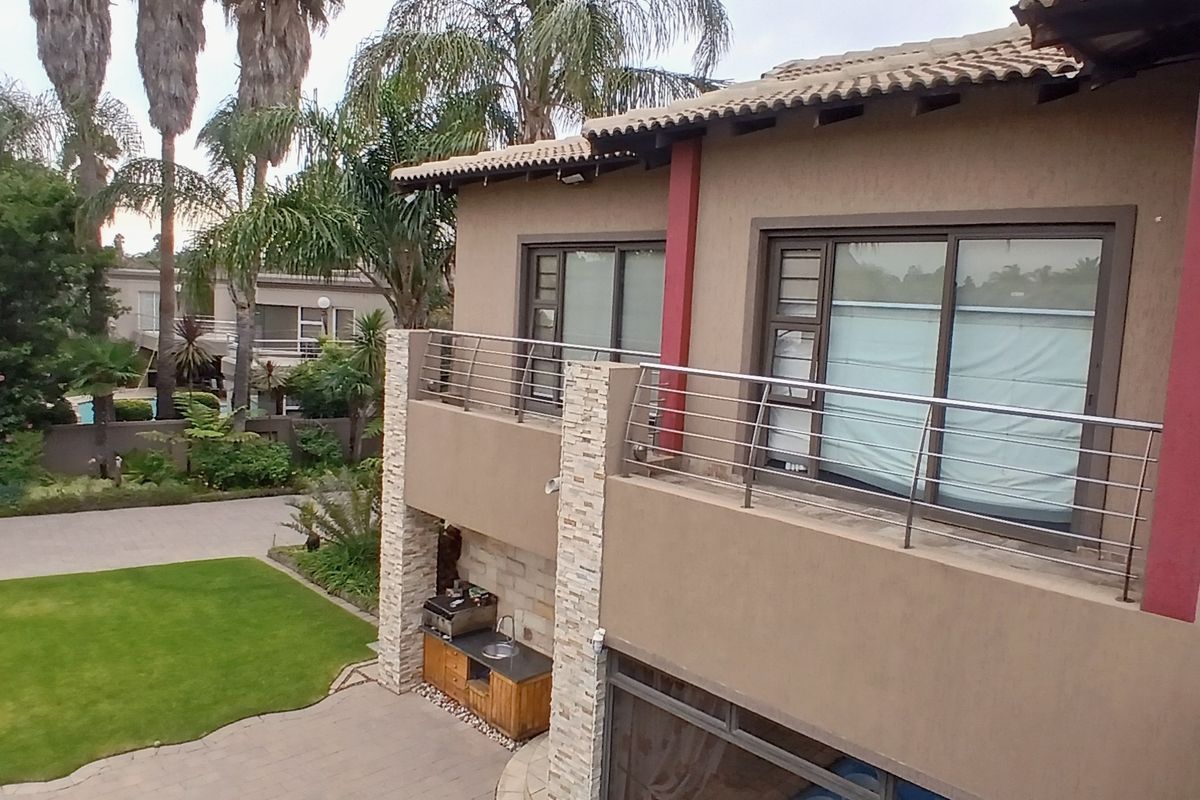 Rear elevation featuring a spacious balcony with stainless steel railings, overlooking a landscaped garden and a paved patio area with a built-in braai.