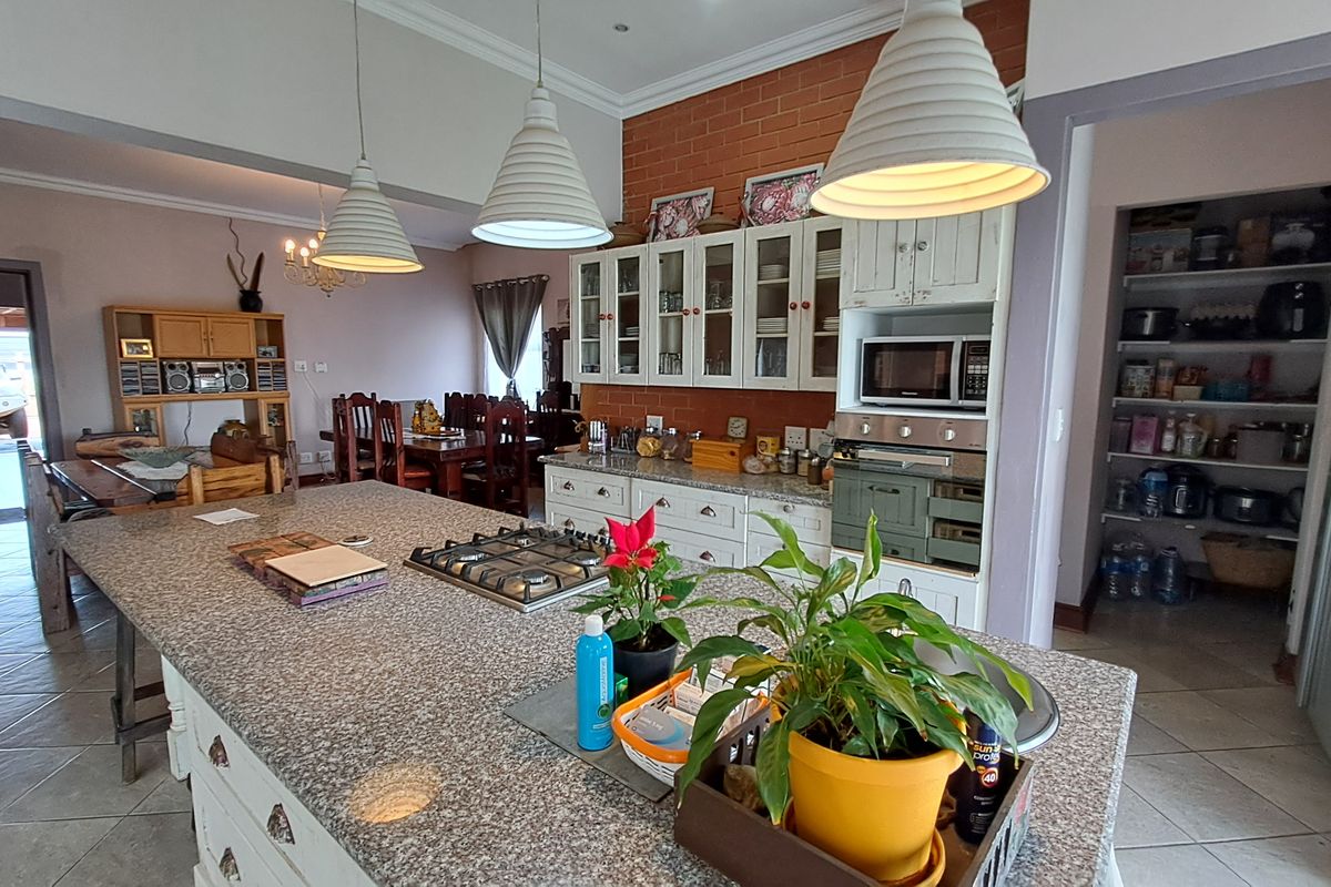 Spacious kitchen featuring a large granite-topped island with gas hob, built-in oven, glass-front cabinetry, and an exposed brick feature wall, adjacent to a walk-in scullery.