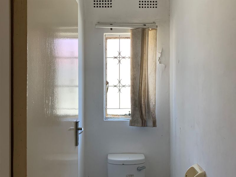 A small, dated guest toilet featuring a standard white toilet suite and a window with security bars, requiring cosmetic updates and wall repairs.
