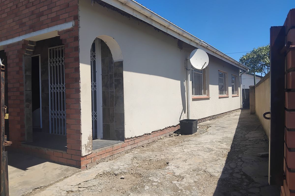 Side view of the property featuring a concrete driveway, plastered exterior walls, and security gates, showing signs of wear and maintenance requirements.