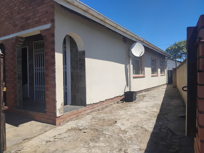 Side view of the property featuring a concrete driveway, plastered exterior walls, and security gates, showing signs of wear and maintenance requirements.
