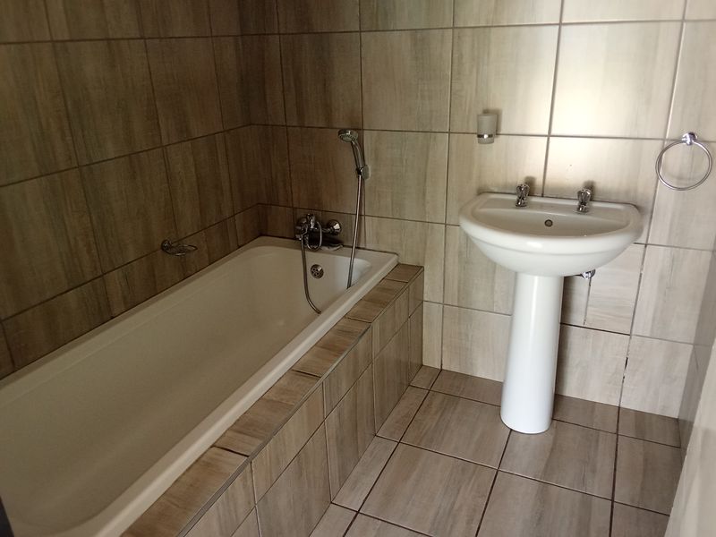 A functional bathroom featuring a built-in bathtub with shower mixer and a pedestal washbasin, finished with neutral ceramic wall and floor tiling.