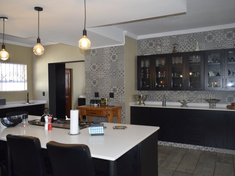 Modern kitchen featuring a large central island with quartz countertops, dark cabinetry, glass-fronted display units, and a decorative tiled feature wall.