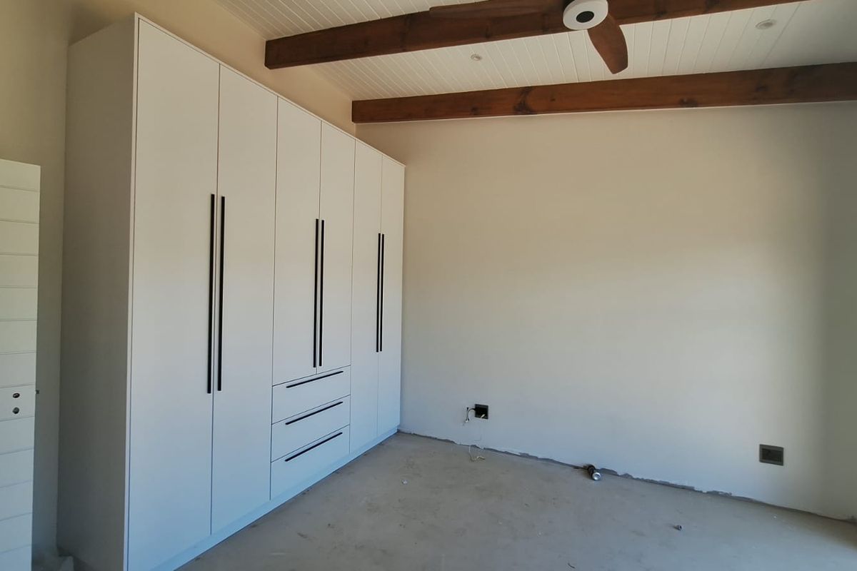 A bedroom featuring modern built-in cupboards, exposed timber ceiling beams, and a ceiling fan, currently awaiting final flooring installation.