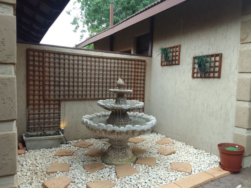 A tranquil garden courtyard featuring a tiered stone fountain, decorative wall trellises, and pebble landscaping set against a textured exterior wall.