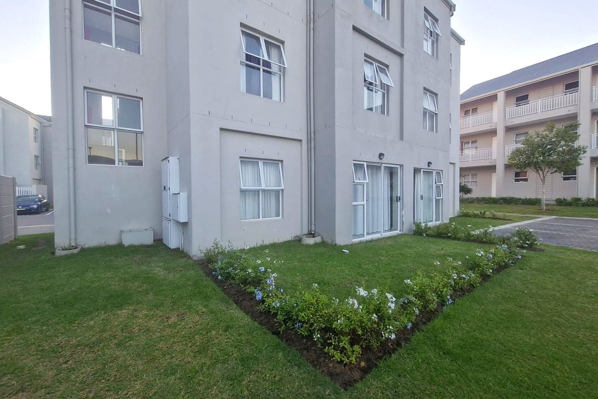 Exterior view of the apartment building featuring a well-maintained lawn, landscaped garden beds, and sliding glass doors providing access to the unit.