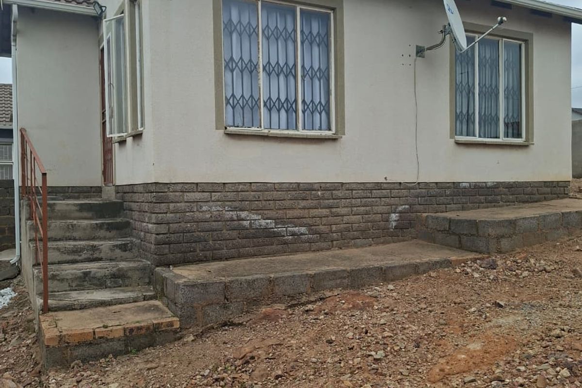 Rear exterior view of a single-story house featuring a brick plinth, security-barred windows, and concrete access steps with metal railings.
