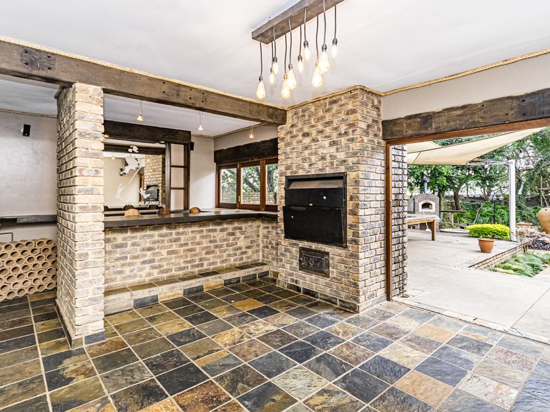 Covered outdoor entertainment area featuring a built-in braai, exposed timber beams, slate flooring, and a convenient prep sink, opening onto the garden.
