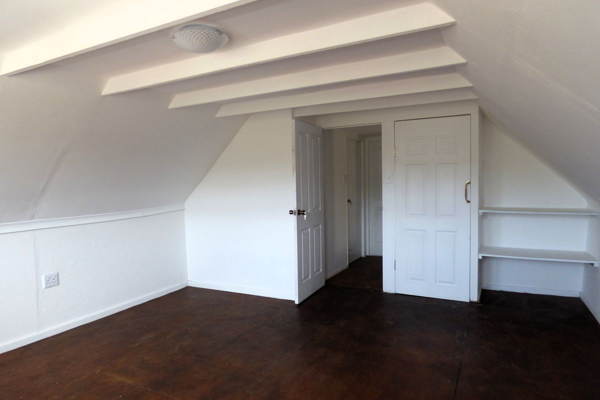 An empty attic-style bedroom featuring sloped ceilings, exposed beams, built-in shelving, and panelled doors, requiring cosmetic updates to the flooring and wall finishes.