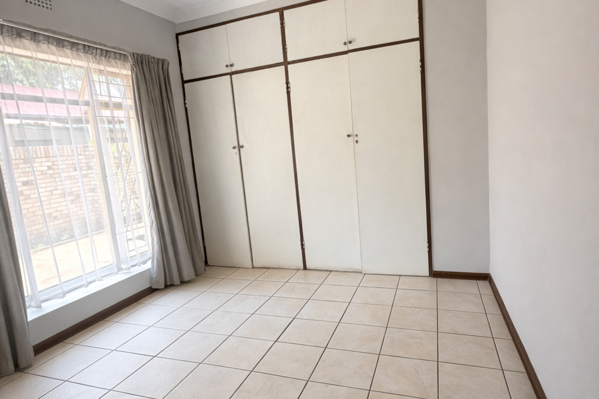 A clean, empty bedroom featuring neutral tiled flooring, built-in storage cupboards, and a window fitted with security bars.