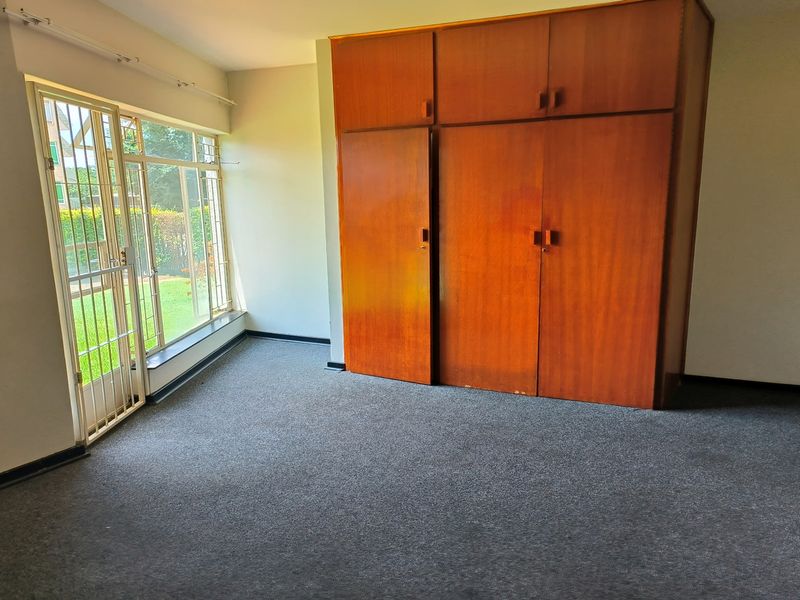 A bedroom featuring built-in wooden cupboards, grey carpeting, and a large window with security bars providing natural light and garden views.