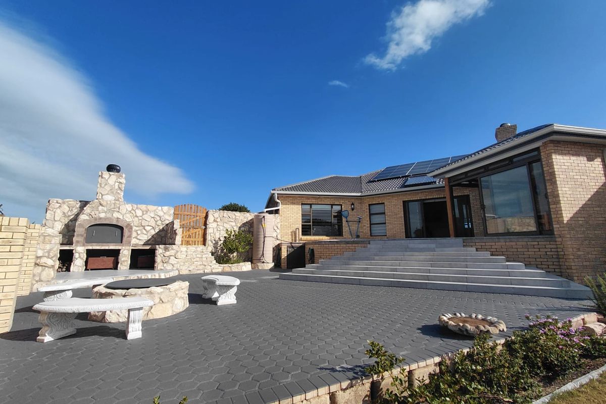 Spacious rear patio featuring a built-in stone braai, paved flooring, and wide concrete steps leading to the main residence with solar panels.