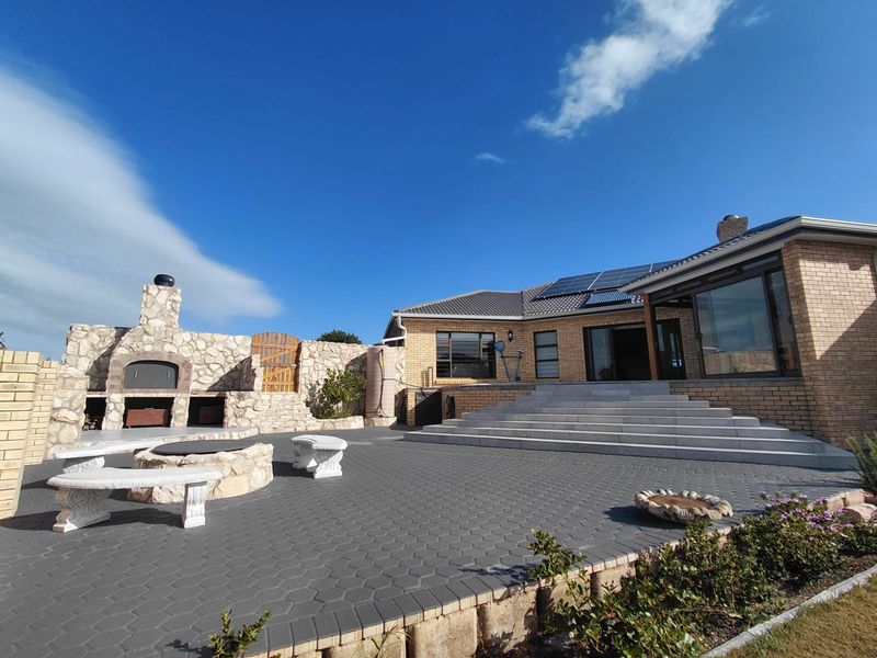 Spacious rear patio featuring a built-in stone braai, paved flooring, and wide concrete steps leading to the main residence with solar panels.
